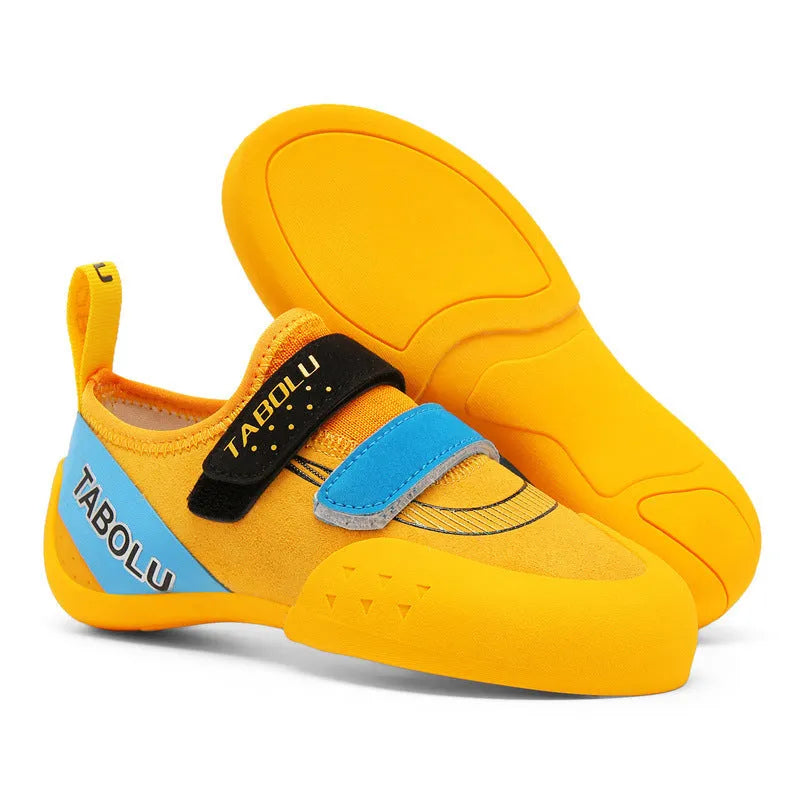 A pair of beginner rock climbing shoes with Velcro straps and a coloured trim, shown on a rocky surface.