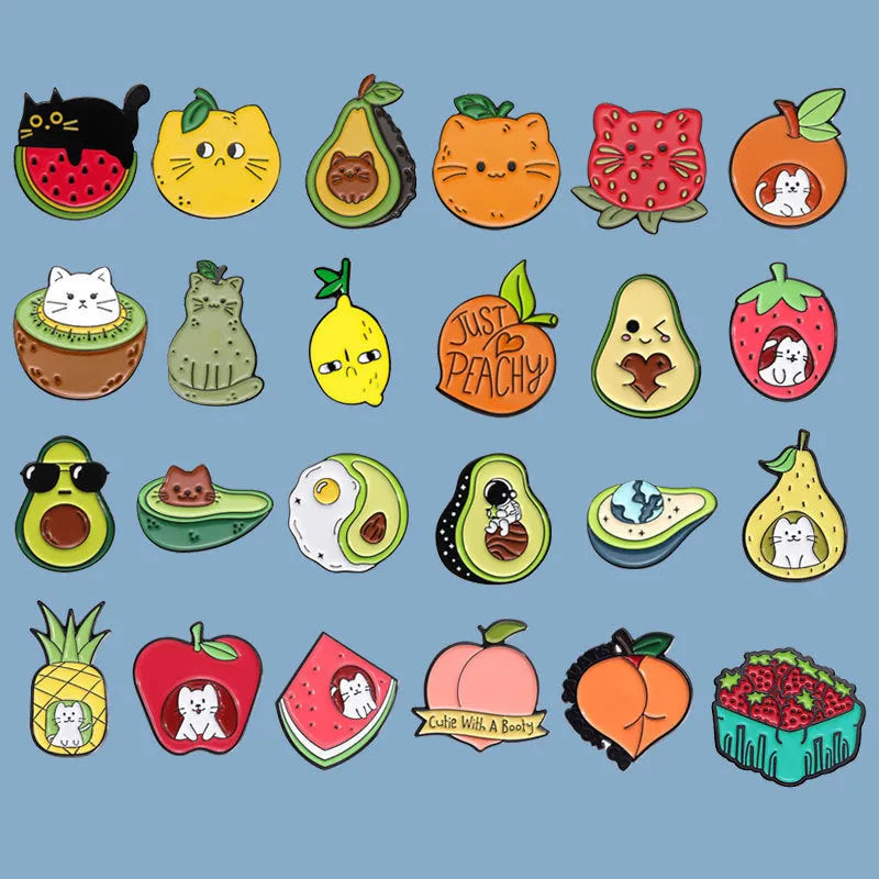 Close-up of a colourful cartoon-style fruit and letter metal brooch with a pin back on a fabric background.