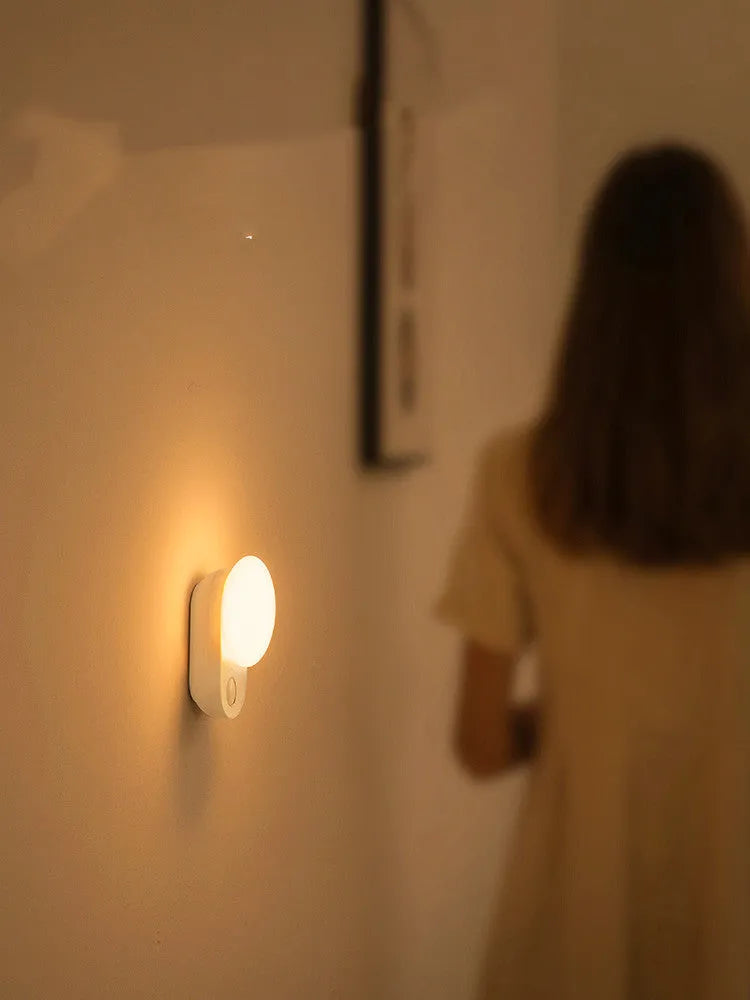 White Baseus rechargeable sensor night light with LED glow, shown on a bedside table.