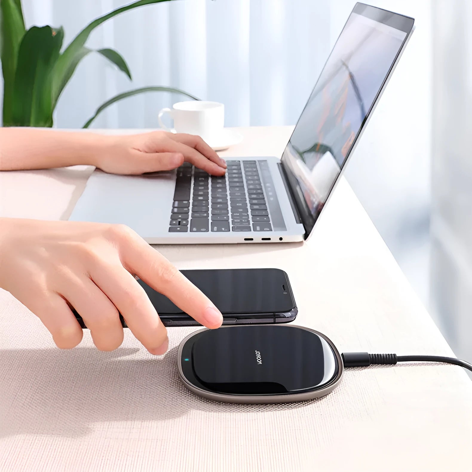 Black square 15W wireless charging pad with glass top on a wooden desk next to a smartphone.