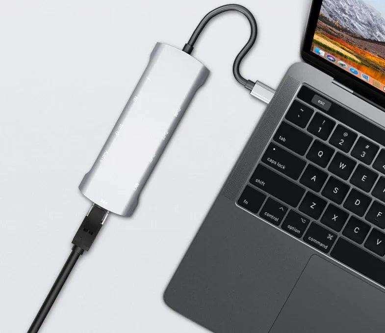 Grey aluminium USB-C docking station with HDMI, VGA, USB, and charging ports on a wooden desk.