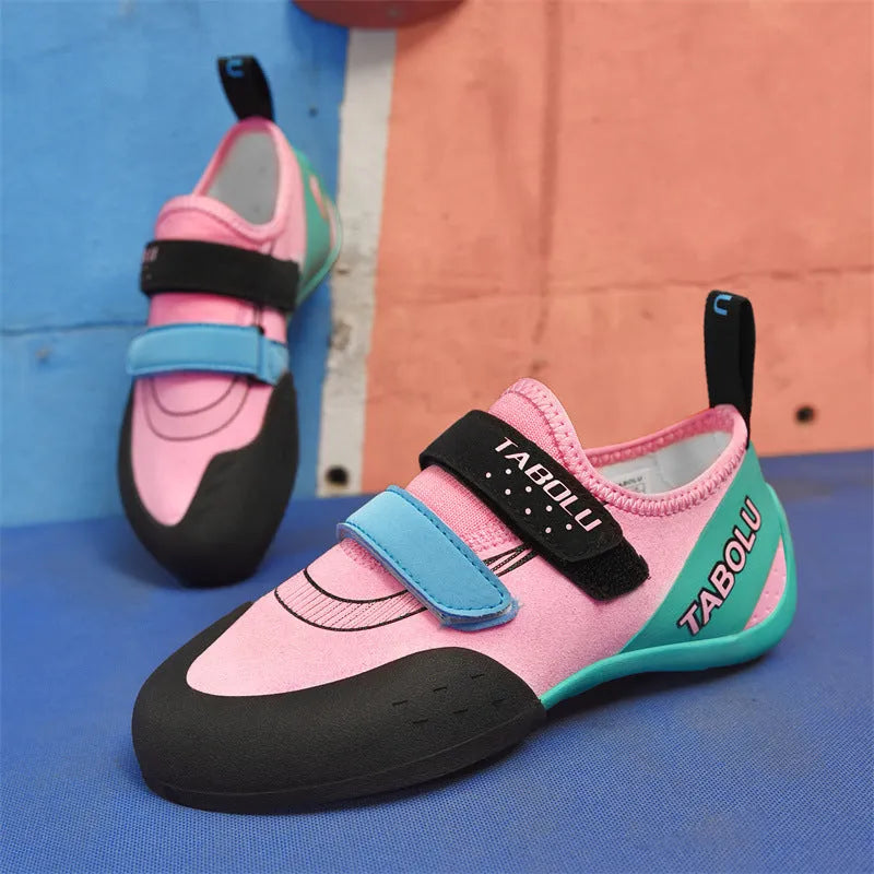 A pair of beginner rock climbing shoes with Velcro straps and a coloured trim, shown on a rocky surface.