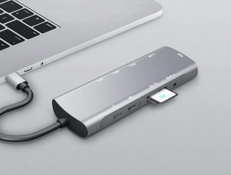 Grey aluminium USB-C docking station with HDMI, VGA, USB, and charging ports on a wooden desk.