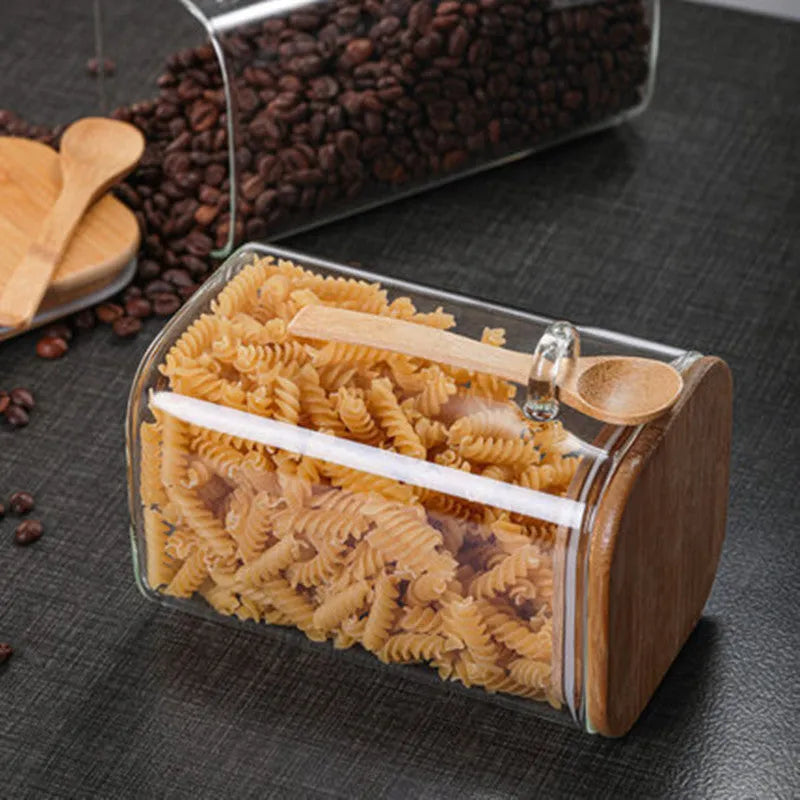 Square glass coffee bean storage jar with wooden spoon and bamboo lid on a kitchen counter