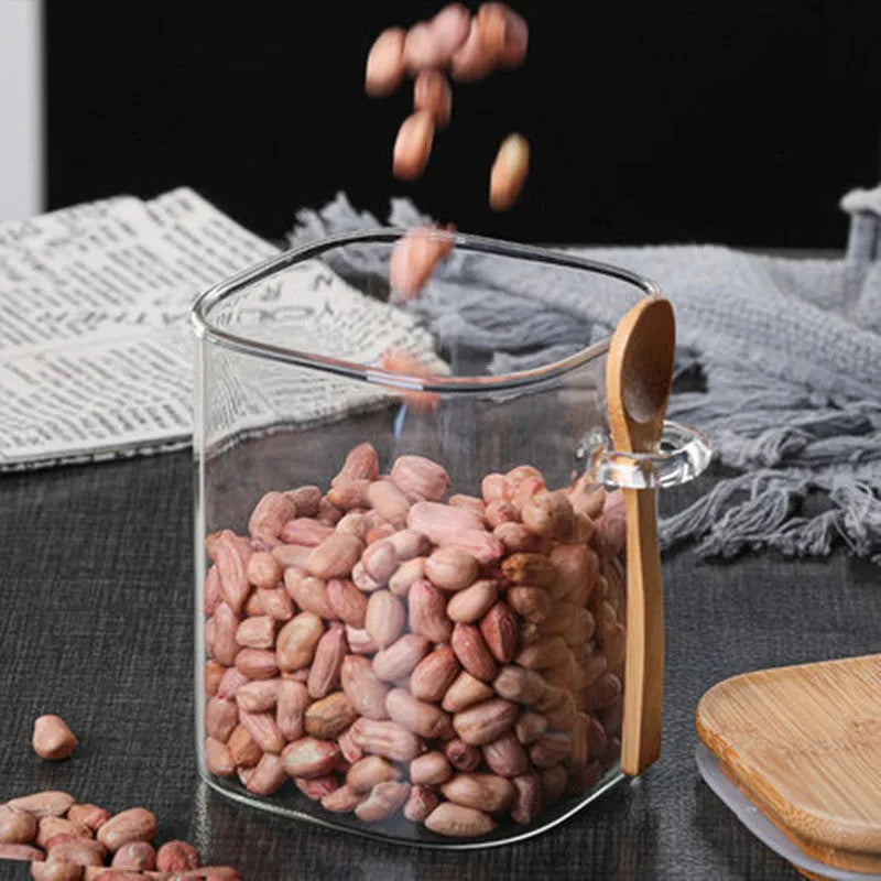 Square glass coffee bean storage jar with wooden spoon and bamboo lid on a kitchen counter