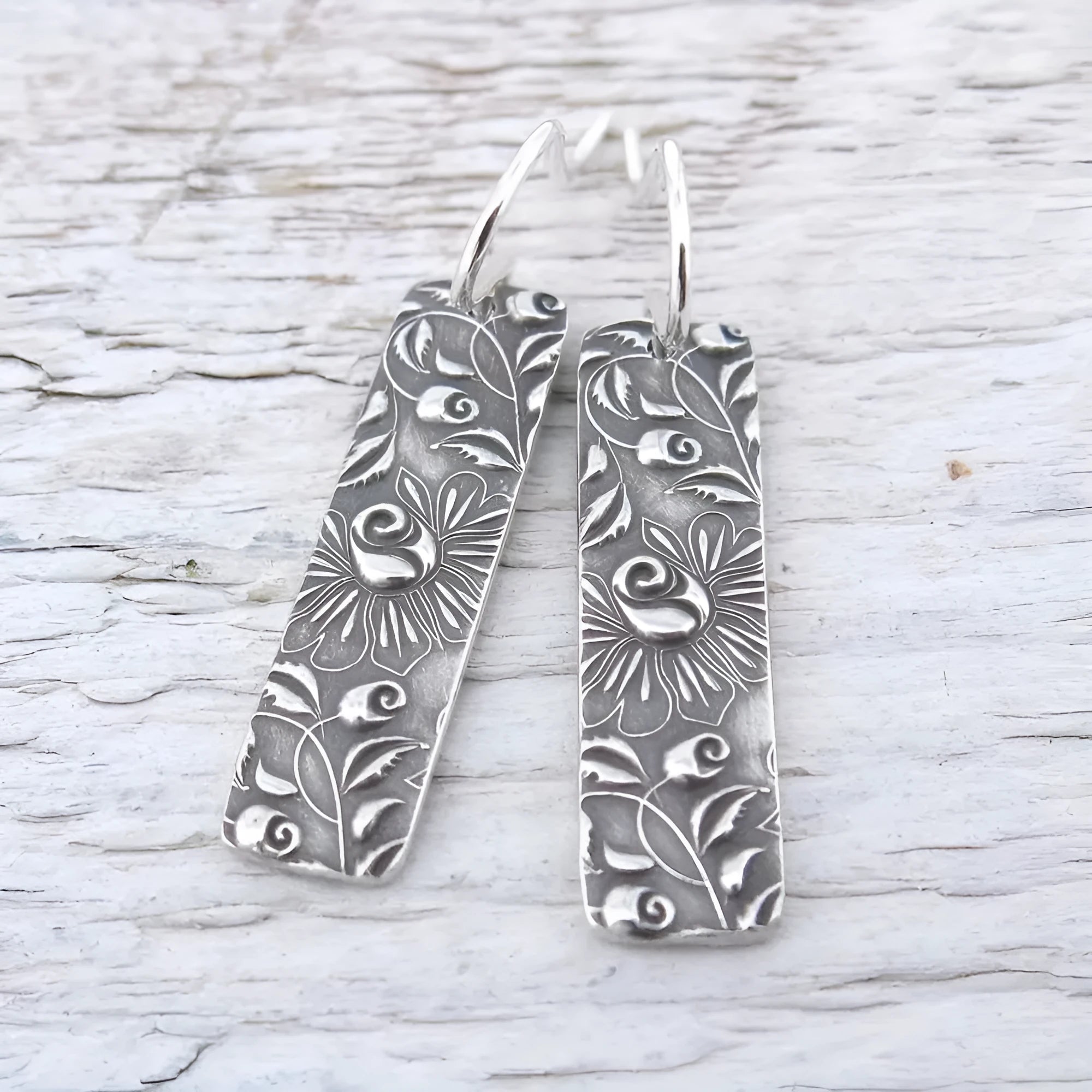 Silver geometric long strip drop earrings with carved vine pattern on a white background.