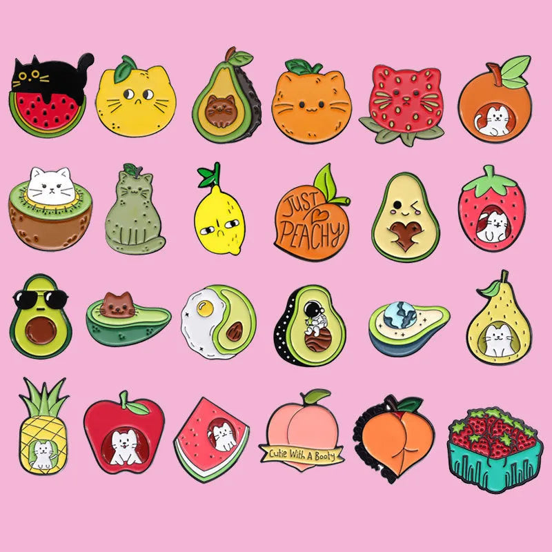 Close-up of a colourful cartoon-style fruit and letter metal brooch with a pin back on a fabric background.