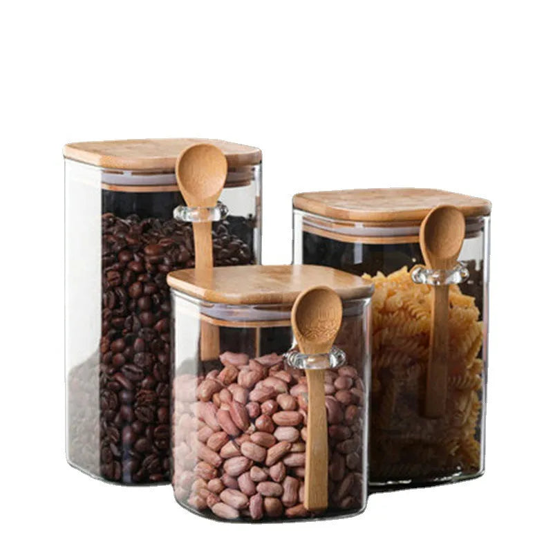 Square glass coffee bean storage jar with wooden spoon and bamboo lid on a kitchen counter