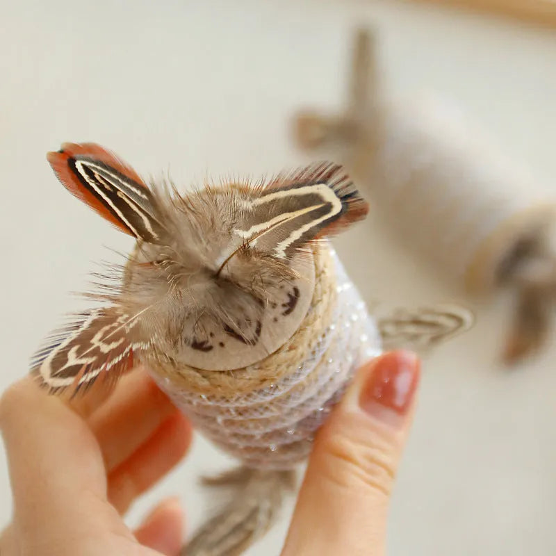 Beige cat toy with hemp rope texture, feathers, and a bell on a wooden roller for scratching and play.