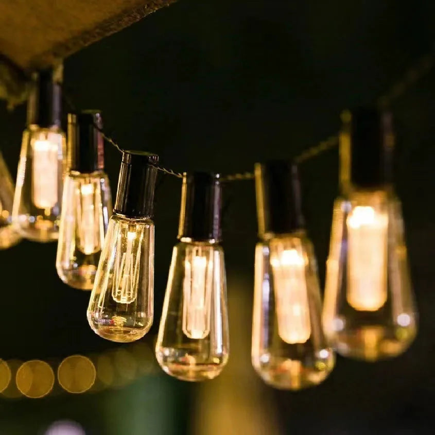 String of retro long infusion bottle shaped outdoor fairy lights hung in a garden.