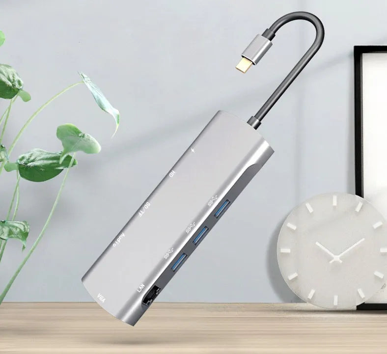 Grey aluminium USB-C docking station with HDMI, VGA, USB, and charging ports on a wooden desk.