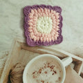 Hand-crocheted wool coaster with lattice pattern in Dream Purple colour on a wooden table.