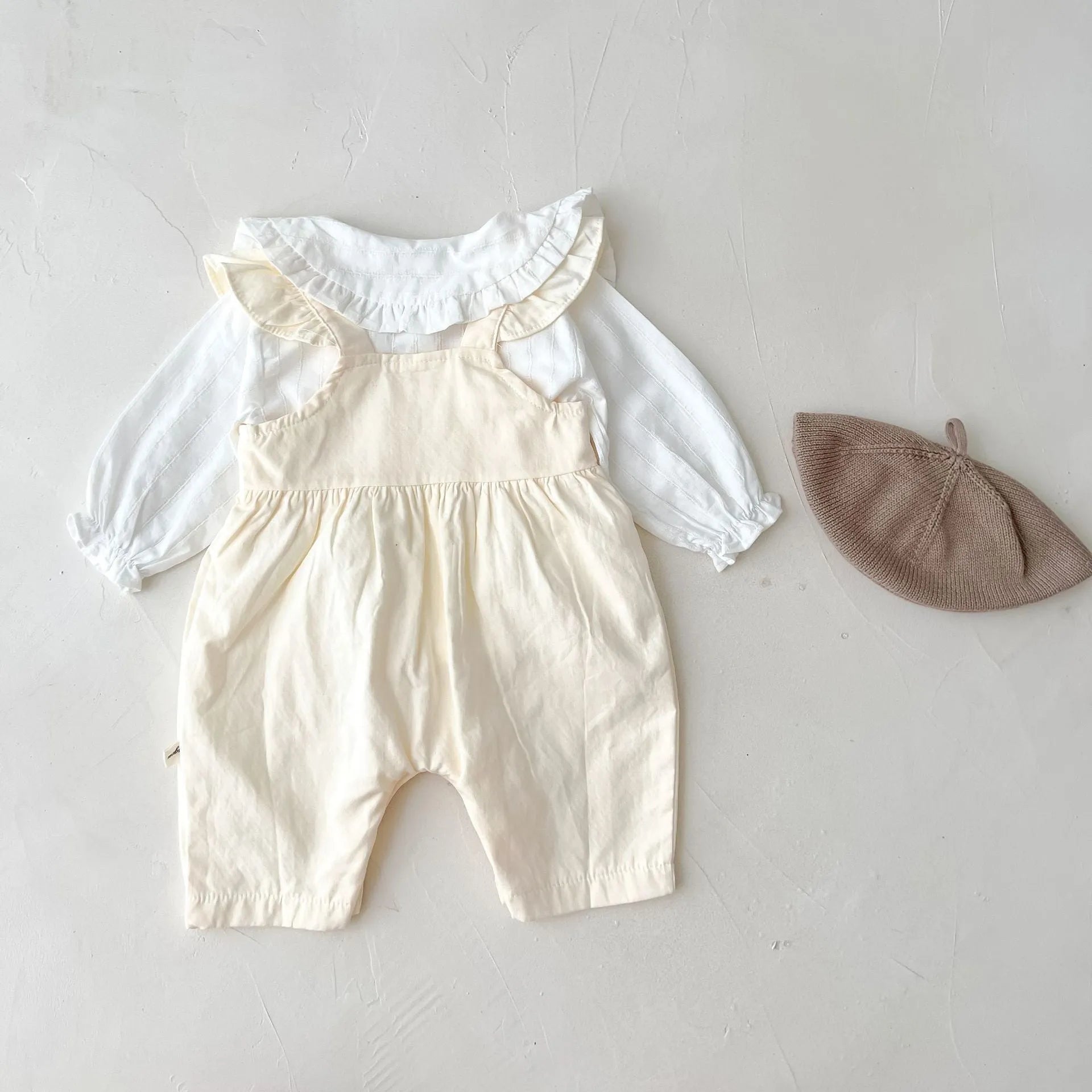 Baby girl wearing a loose-fit cotton suspender romper with adjustable straps in an apricot colour.