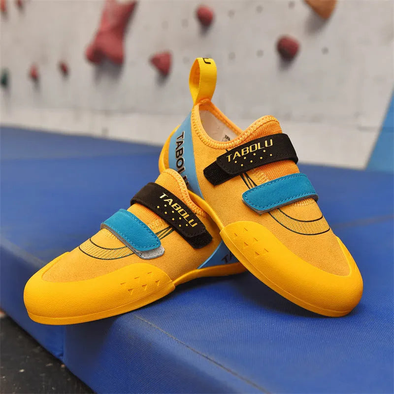 A pair of beginner rock climbing shoes with Velcro straps and a coloured trim, shown on a rocky surface.