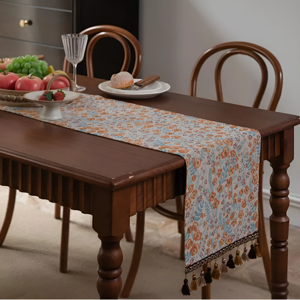 Long beige jacquard table runner with floral pattern and knotted tassels on a wooden dining table.