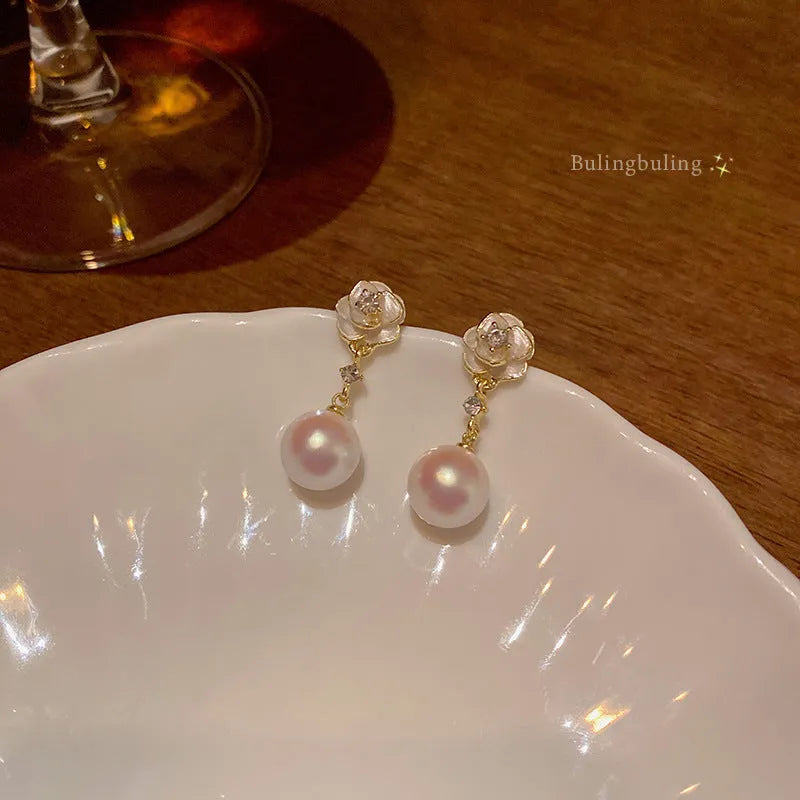 Gold-tone camellia flower stud earrings with a white crystal pearl in the centre on a white background.