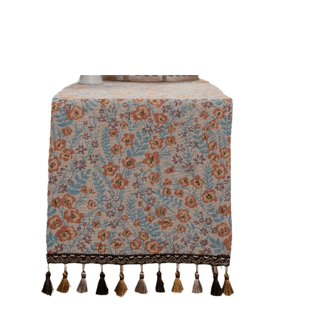 Long beige jacquard table runner with floral pattern and knotted tassels on a wooden dining table.