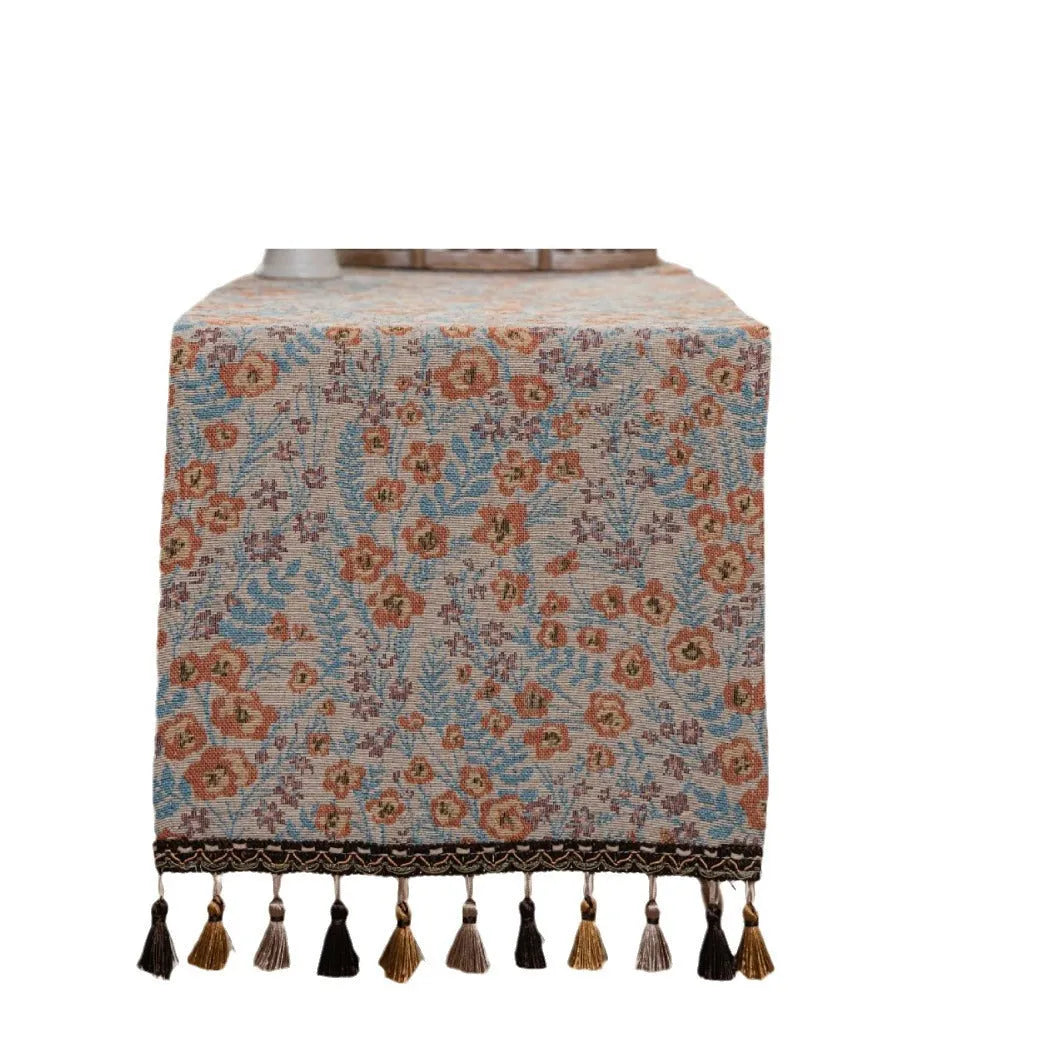 Long beige jacquard table runner with floral pattern and knotted tassels on a wooden dining table.