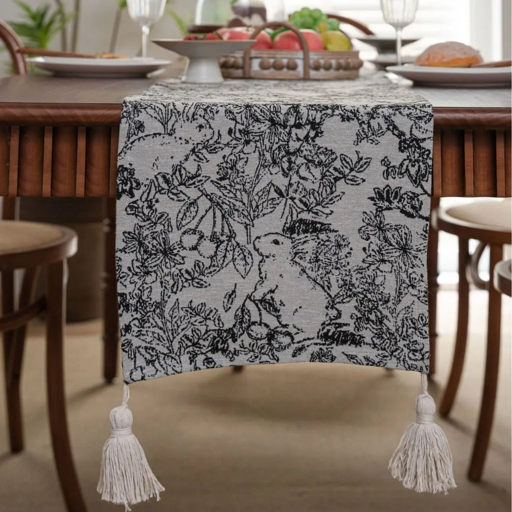 Long beige jacquard table runner with floral pattern and knotted tassels on a wooden dining table.