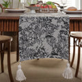 Long beige jacquard table runner with floral pattern and knotted tassels on a wooden dining table.