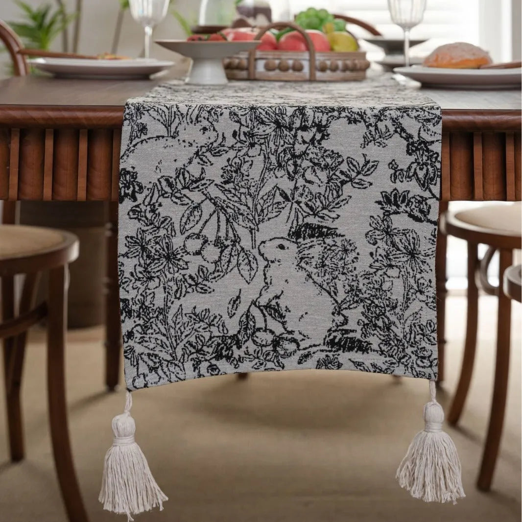 Long beige jacquard table runner with floral pattern and knotted tassels on a wooden dining table.