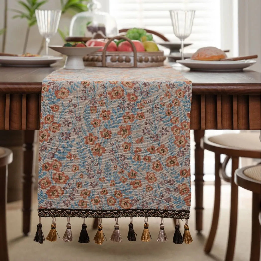 Long beige jacquard table runner with floral pattern and knotted tassels on a wooden dining table.