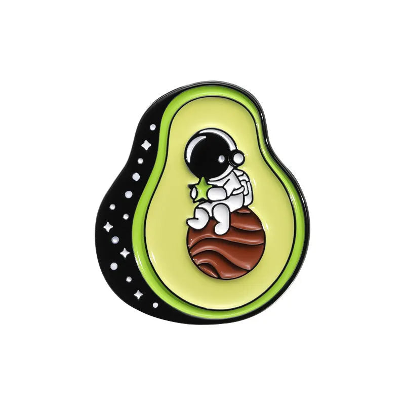 Close-up of a colourful cartoon-style fruit and letter metal brooch with a pin back on a fabric background.