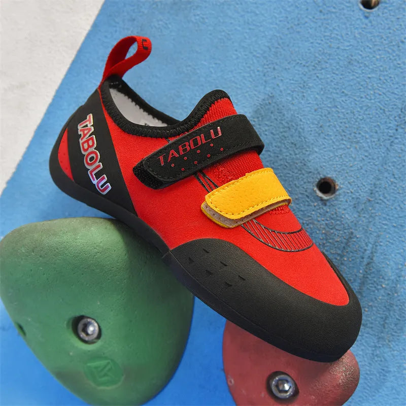 A pair of beginner rock climbing shoes with Velcro straps and a coloured trim, shown on a rocky surface.