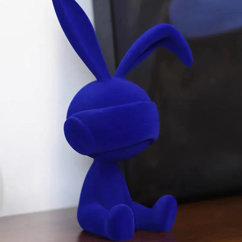 Suede blindfold rabbit resin ornament in blue, green, or red colour on a table.