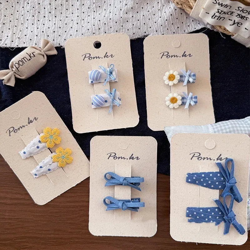 One pair of children's cloth hair clips with bow and flower detailing, shown in a choice of five styles and two clip sizes.