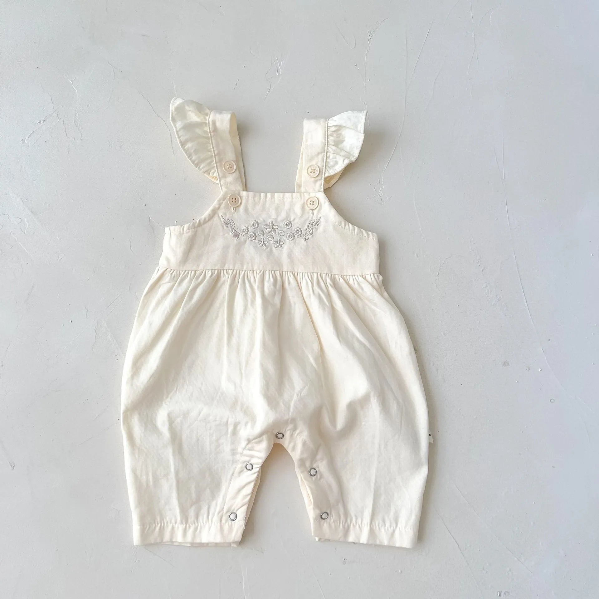 Baby girl wearing a loose-fit cotton suspender romper with adjustable straps in an apricot colour.